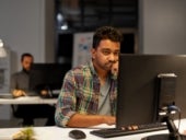 A visibly tired/frustrated man working at a computer in an office setting with another coworker in the background